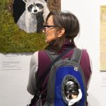 Cricket peeks out from her pack as she rides around with Sonia Murray, from Fairfield, as they look at North American Raccoon a free motion embroidery on canvas by JoAnne Lincoln at The Nature of Thread art quilt show at Mare Island Art Studios on Saturday. (Chris Riley/Times-Herald)